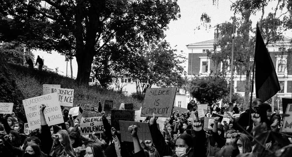 A group of protestors holding banners and parading through a street. The slogans say No Justice, Peace, I understand that i will never understand however I stand. Silence is violence. Silence is complicity. To be silent is to be complicit. Black Lives Matter.