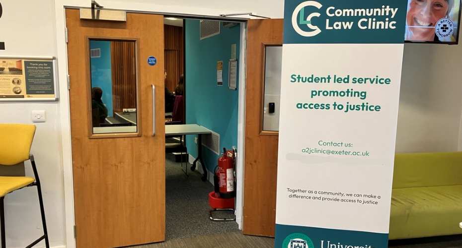 A pull up sign displaying the Community Law Clinic logo. Student led service promoting access to justice. Contact us: a2jclinic@exeter.ac.uk, Together as a community, we can make a difference and provide access to justice. It is stood in front of an open door to the room called Rougemont.