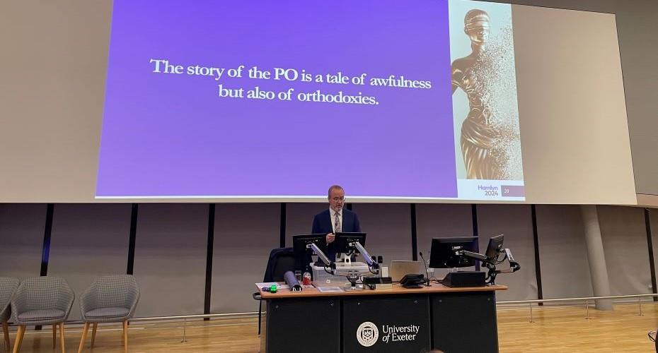 A large screen displaying the words The story of the PO is a tale of awfulness but also of orthodoxies. The Hamlyn Lectures 2024. There is a person, professor Moorhead stood on stage in front of the screen, with a large lectern which has computer screens and microphones