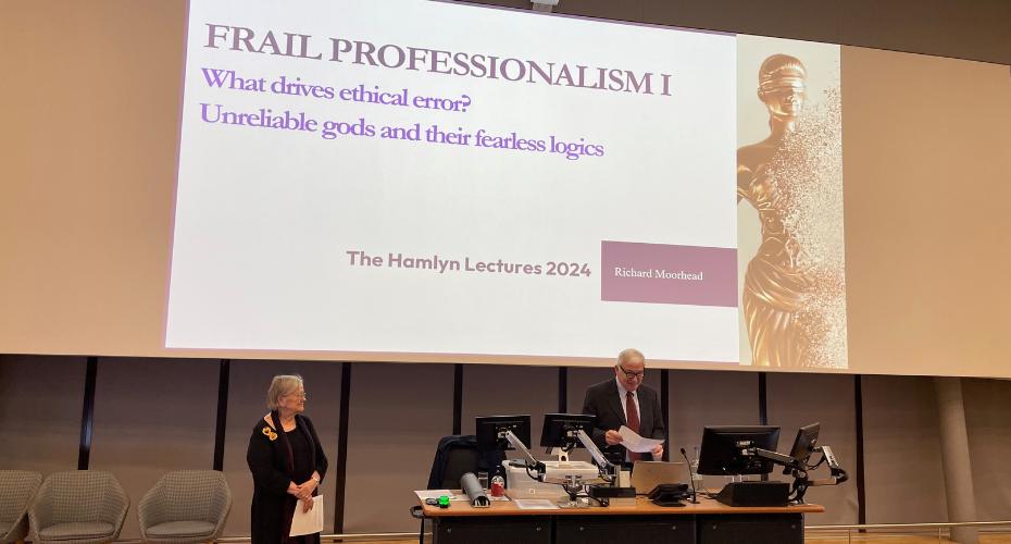 A large screen displaying the words Frail Professionalism, What drives ethical error? Unreliable gods and their fearless logics.There are two people on stage in front of the screen, with a large lectern which has computer screens and microphones